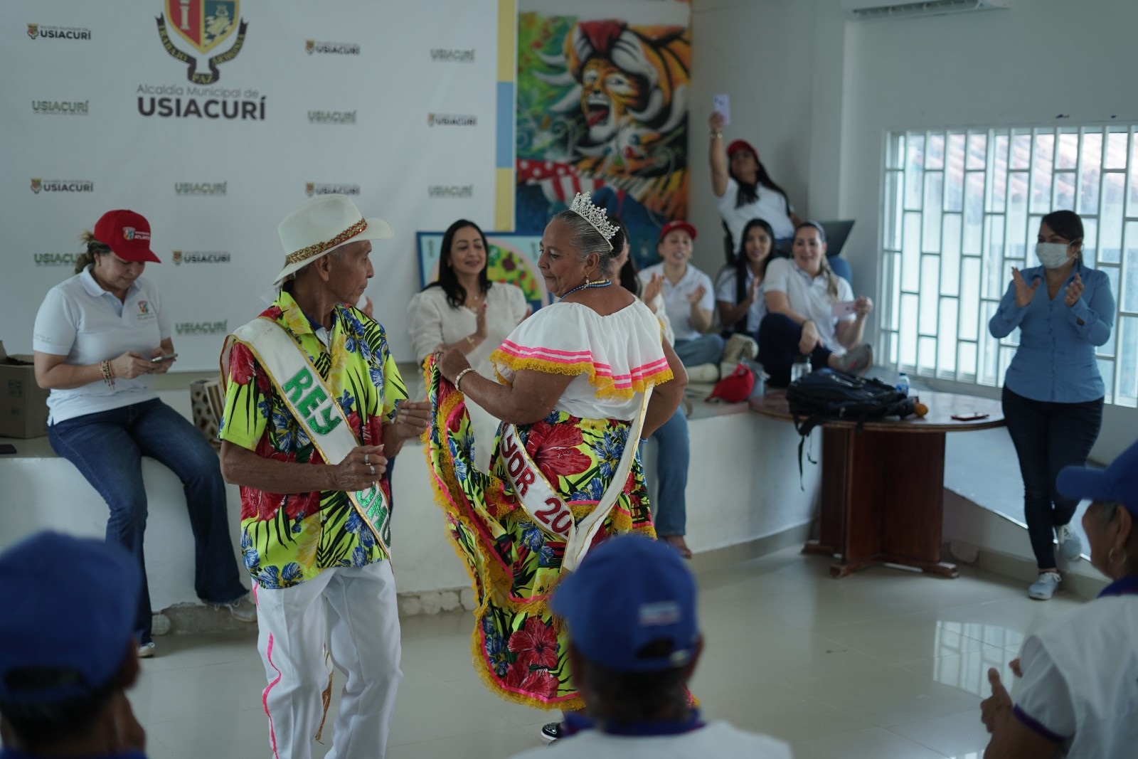 Foto del Rey Momo y la Reina 2024 de los Adultos Mayores bailando frente a otras personas, entre estas, la Gerente de Capital Social, Karina Llanos